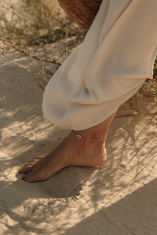 Chaîne de cheville dorée portée sur un pied dans le sable, pour un article sur les chaînes de chevilles et leur signification.